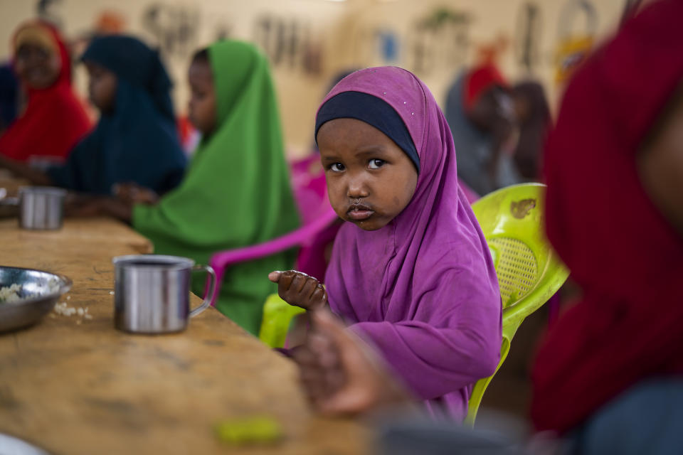 A young girl wearing a purple shawl over her head, seated in front of a metal cup at a trestle table, looks warily at the camera.