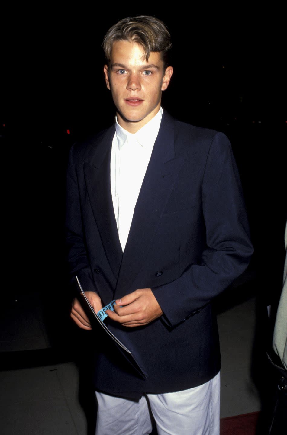 matt damon looks at the camera with a neutral expression on his face, he holds a booklet and wears a navy blue jacket and white collared shirt