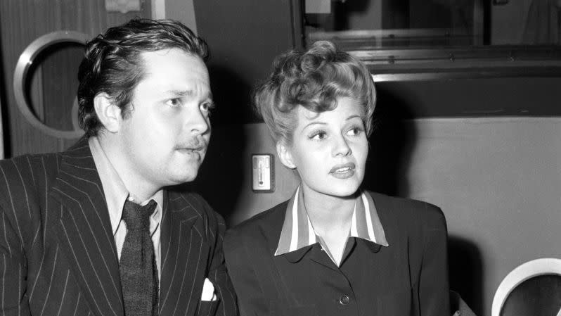orson welles and rita hayworth sit next to each other and look to the right, he wears a dark pinstriped suit jacket with a collared shirt, tie, and gray suit pants, she wears all black with a striped collar visible under her jacket