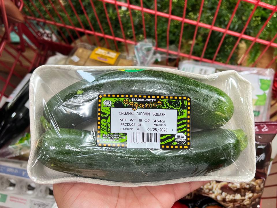 hand holding package with two zucchinis above a cart at trader joes