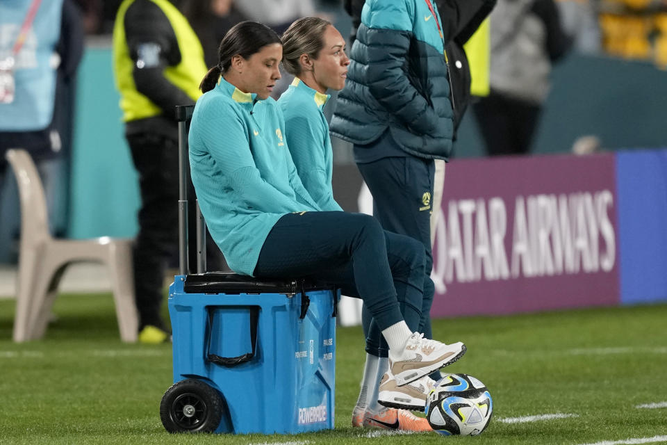 Australia's Sam Kerr, left, sits watching her teammates during warmup before the Women's World Cup soccer match between Australia and Ireland at Stadium Australia in Sydney, Australia, Thursday, July 20, 2023. Australia's team captain Kerr will miss their her team's World Cup opening match due to an injury. (AP Photo/Mark Baker)