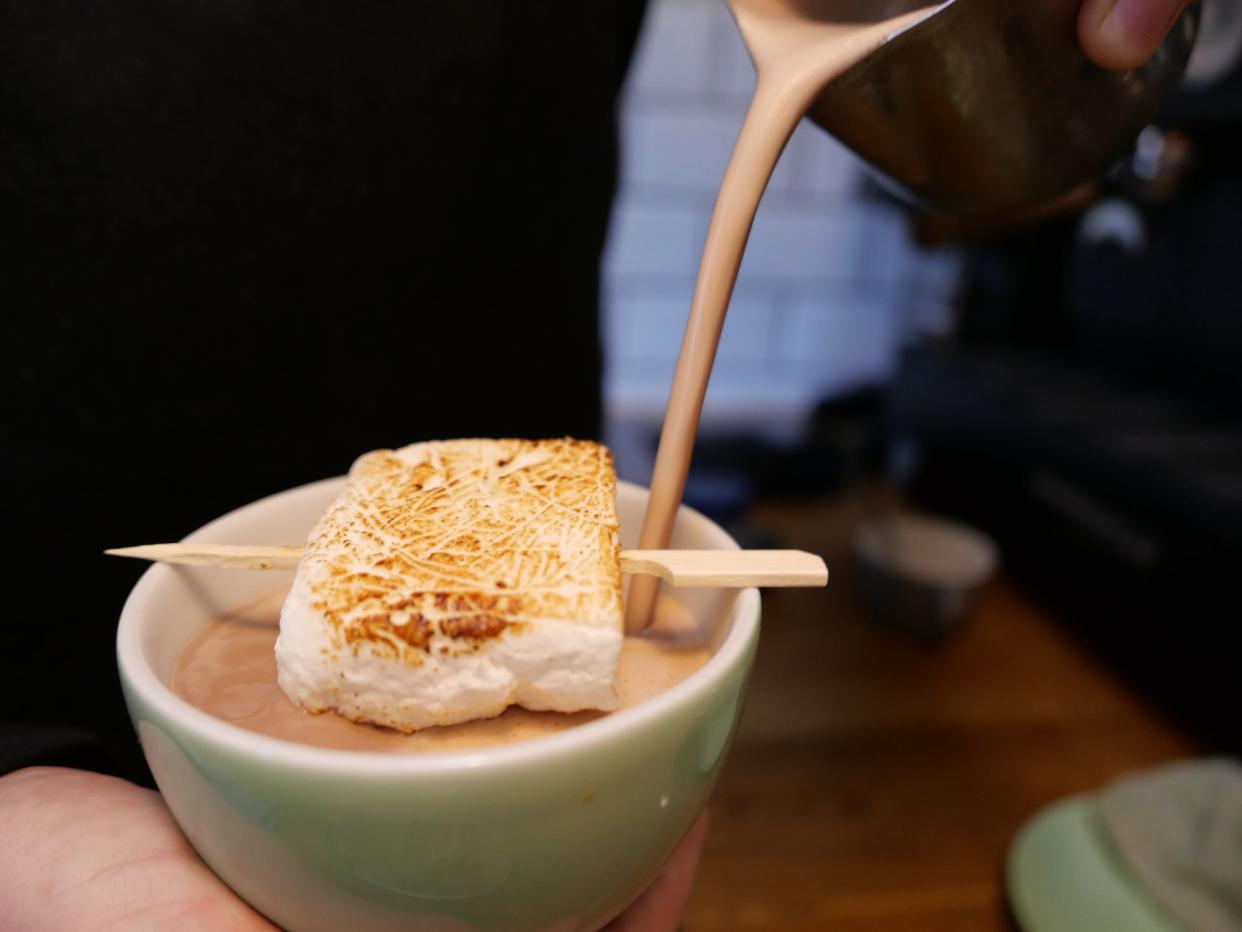 Hot chocolate being poured into a china cup, with a toasted marshmallow on a skewer lying across it. 