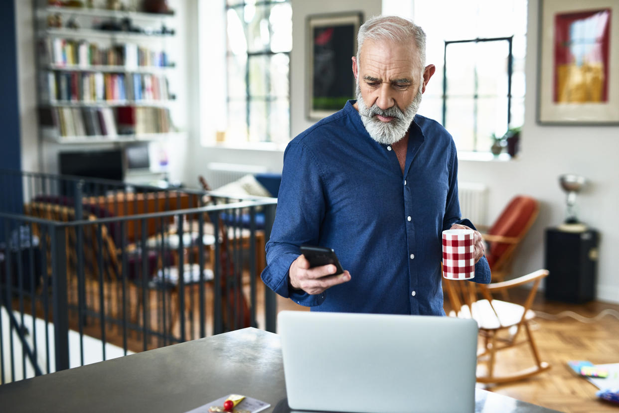 Man in his 50s in apartment, texting, with laptop on work surface, serious expression on face, planning for the day ahead, drinking coffee