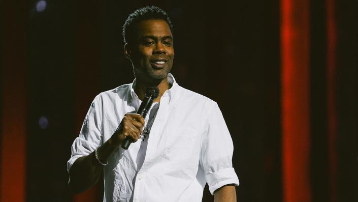 Chris Rock walks on the stage for his Netflix standup special holding a microphone wearing a white shirt