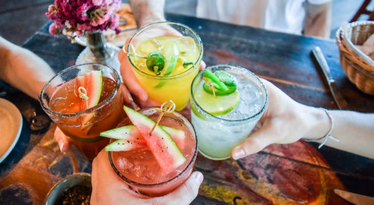 A photo of four people holding glasses with cocktails in them out in a toast, over a wooden table.