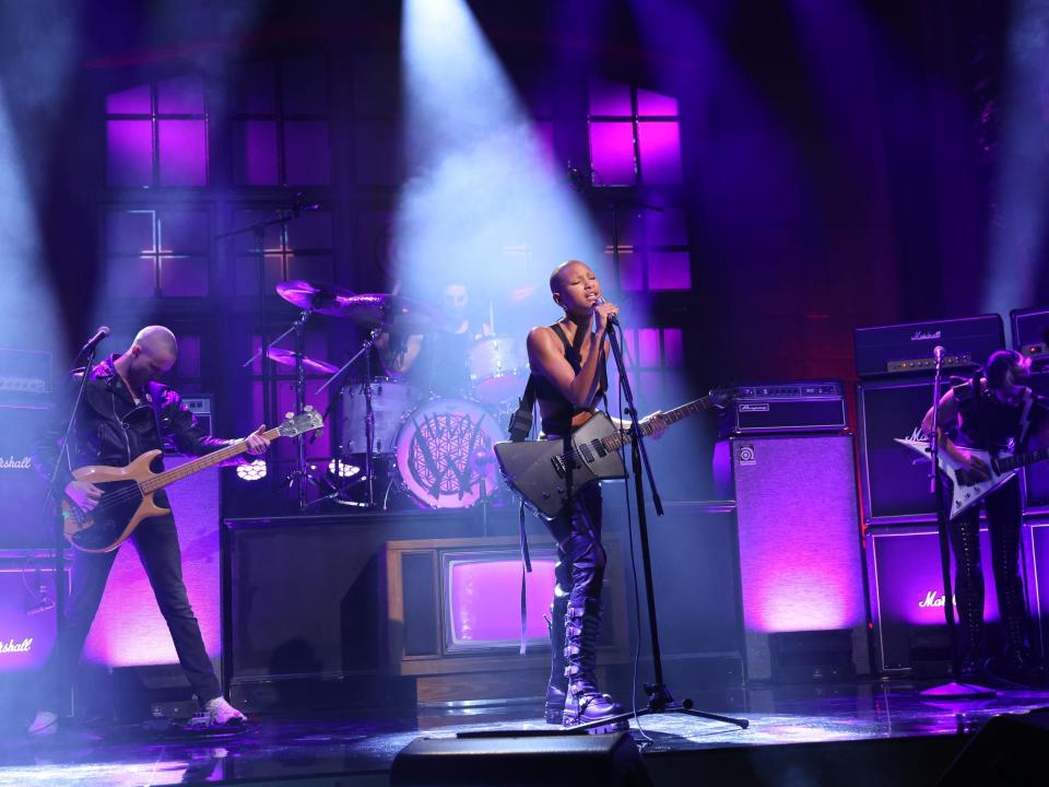 Willow performs on stage surrounded by items underlit with purple lighting. She's in the smoky spotlight, one hand holding the mic stand, guitar hanging from her body.