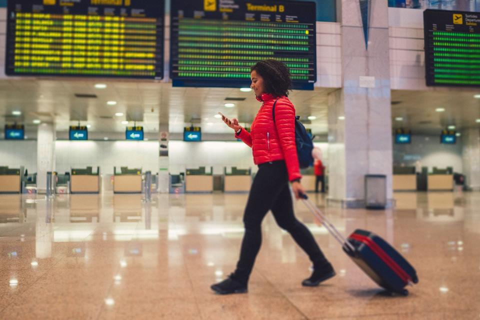 A woman with wheeled luggage walking at the airport and texting without a facemask