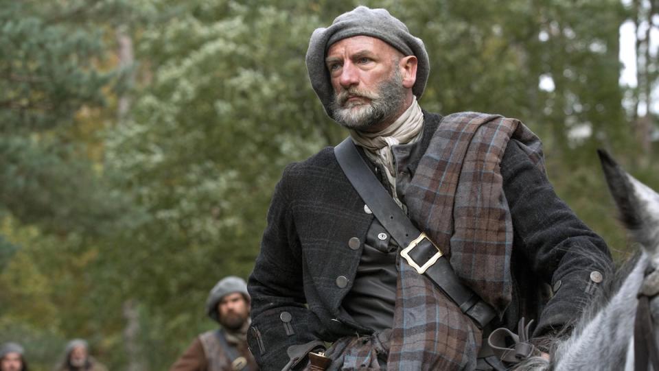 A man in traditional Scottish clothes sits on a horse in the woods