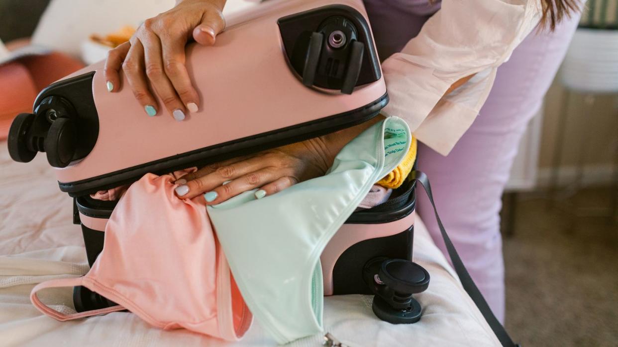 woman with pastel colored nail polish stuffing pink suitcase with swimsuit and clothes