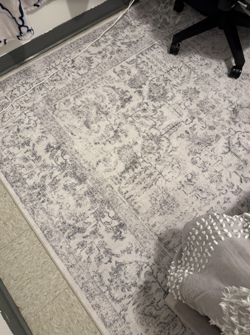 An ornate rug with floral patterns is partially covered by a tasseled blanket. A chair with wheels is visible in the top right corner