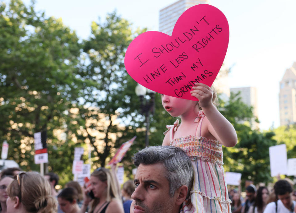 little girl sitting on her father's shoulders holding the sign that says "I shouldn't have less rights than my grandmas"