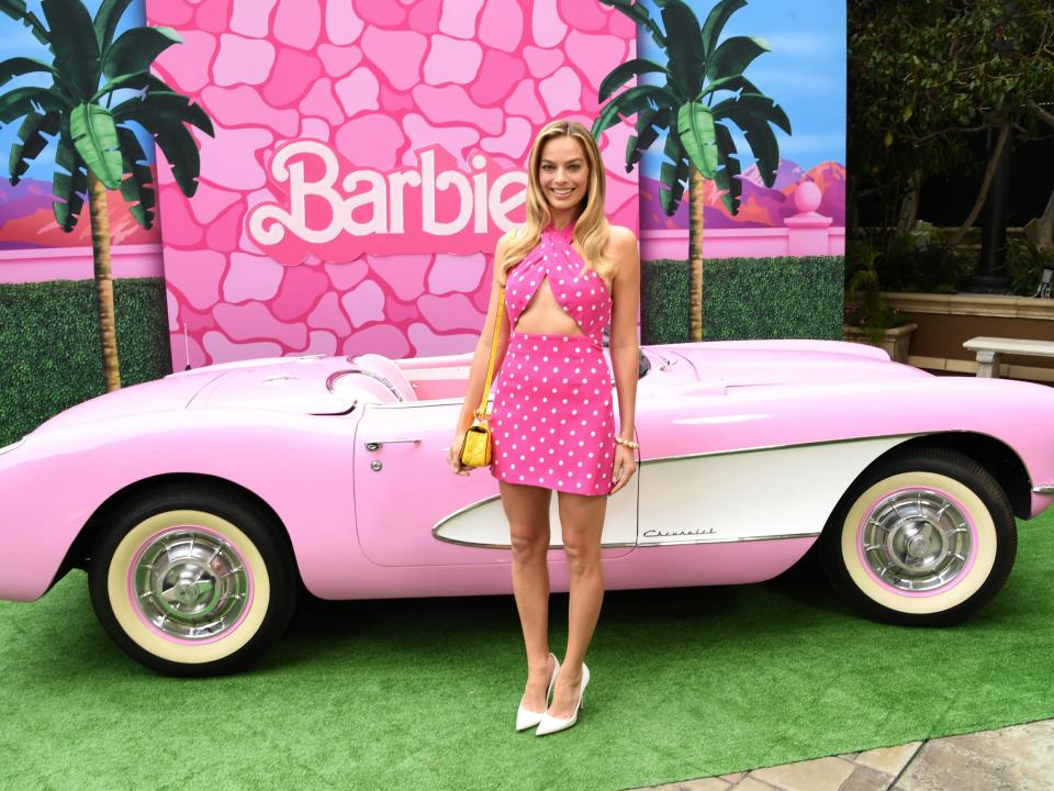 Margot Robbie standing in front of a pink 1956 Chevy while wearing a sleeveless pink dress with white spots.