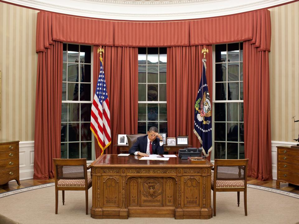 President Barack Obama sits in the Oval Office, which has red curtains and striped wallpaper