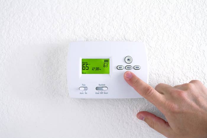 A hand adjusting a wall-mounted digital thermostat, which displays 65°F with system settings for cool and heat