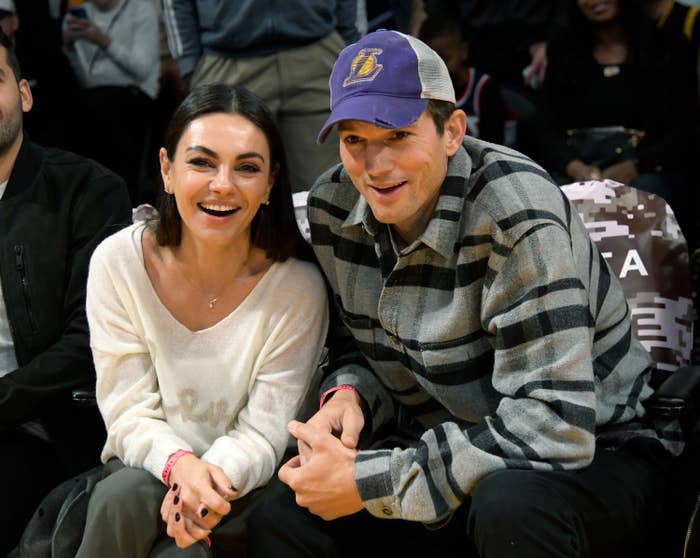 Mila Kunis and Ashton Kutcher sitting courtside at a basketball game, smiling and holding hands. Mila is wearing a white top, Ashton a plaid shirt and baseball cap