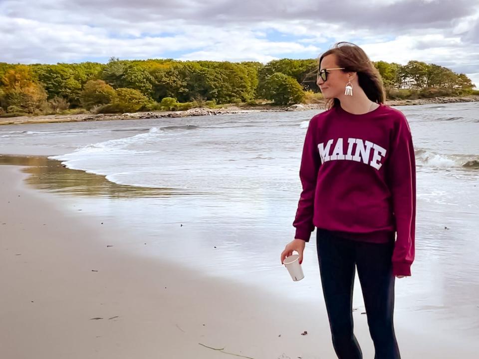 Emily, wearing a maroon sweatshirt with white lettering that reads "Maine," walks along a beach while holding a coffee cup. There are lots of trees in the distance. 