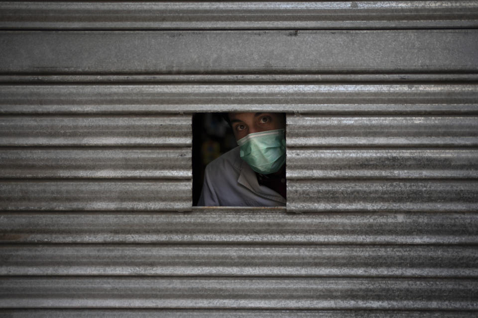 Un hombre con una mascarilla se asoma por una pequeña apertura en la puerta de una farmacia en espera de clientes en Igualada, España, el lunes 6 de abril de 2020. (AP Foto/Felipe Dana)