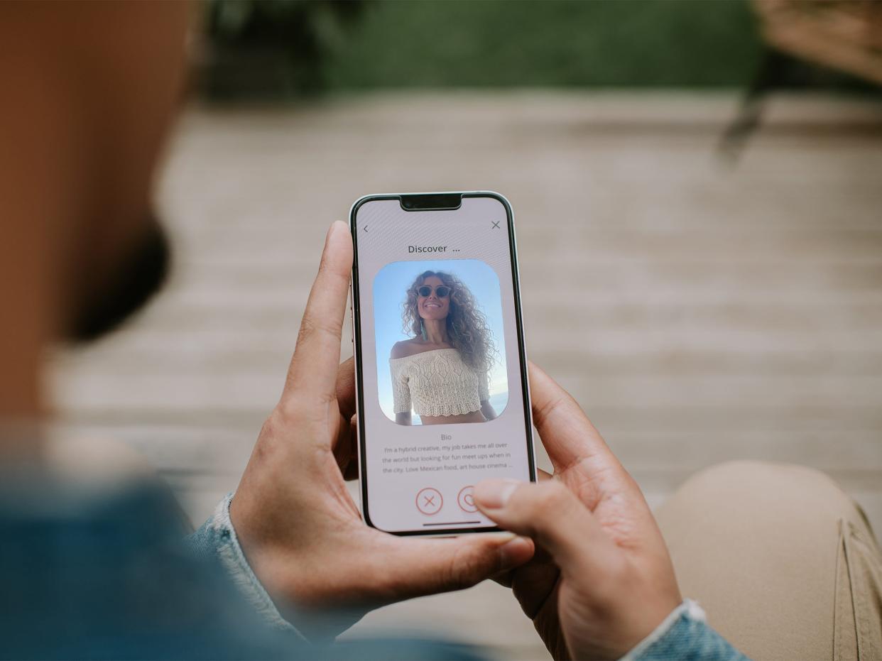 A person using their cellphone to look at a profile on a dating site. The profile shows a woman wearing a crop top and sunglasses