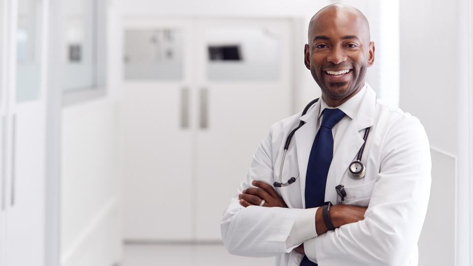 Portrait Of Mature Male Doctor Wearing White Coat Standing In Hospital Corridor.