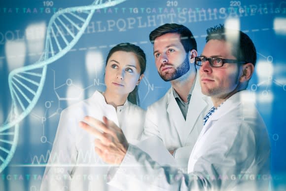 Scientists in lab coats confer in front of a monitor displaying data and a double helix.