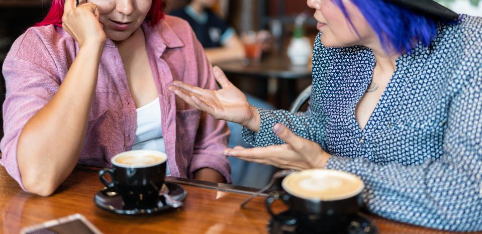 Two individuals are seated at a café table with coffee cups. One appears distressed while the other gestures animatedly, as if offering comfort or advice. Names unknown