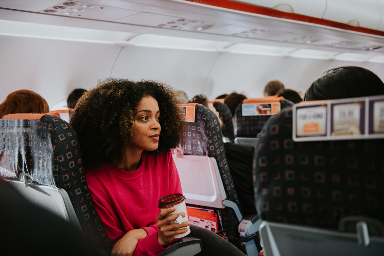 A young woman with a fear of flying sits on a crowded plane on an aisle seat. She clutches a cup of coffee and looks perplexed and thoughtful.