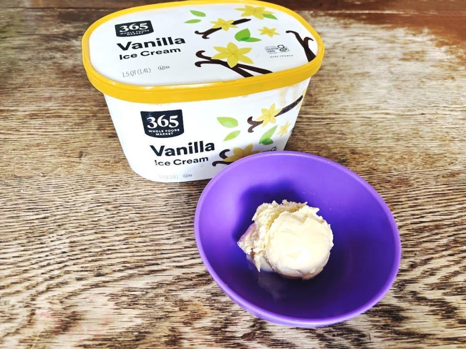 A white carton of 365 vanilla ice cream with a yellow band around the lid sitting next to a purple bowl with a scoop of ice cream in it