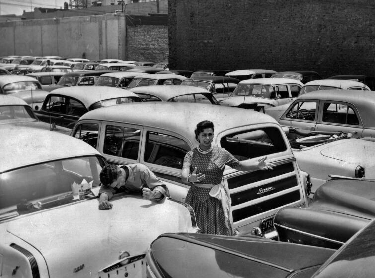 A black and white 1950s photo shows a woman in a dotted dress standing in the middle of a large, overstuffed parking lot
