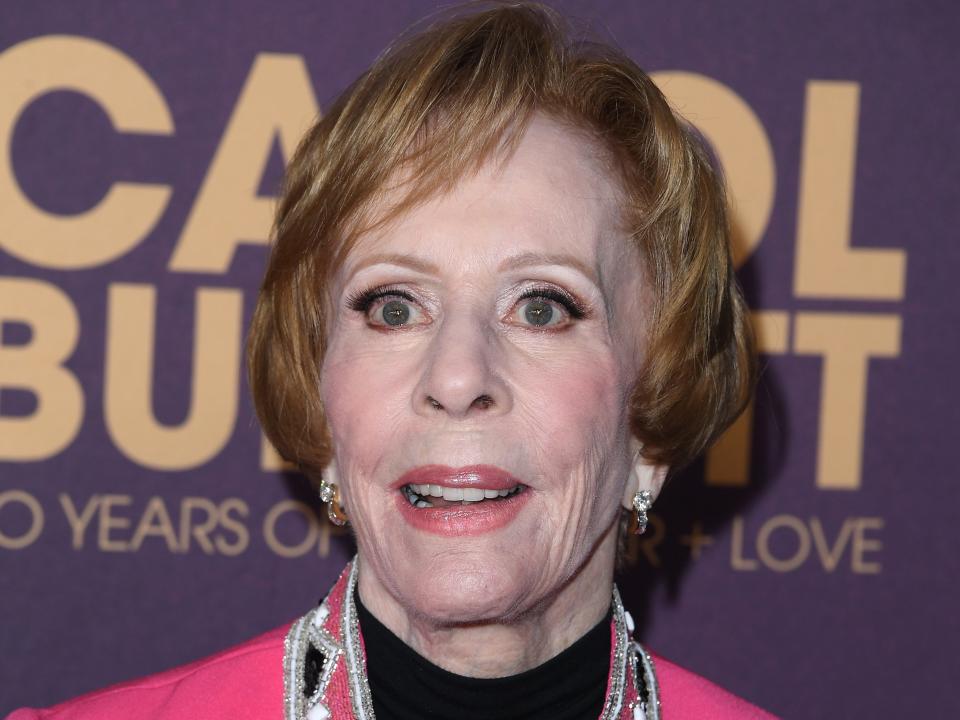 carol burnett in. apink jacket and black shirt, standing in front of a red carpet sign for her "carol burnett: 90 years of laughter and love" special