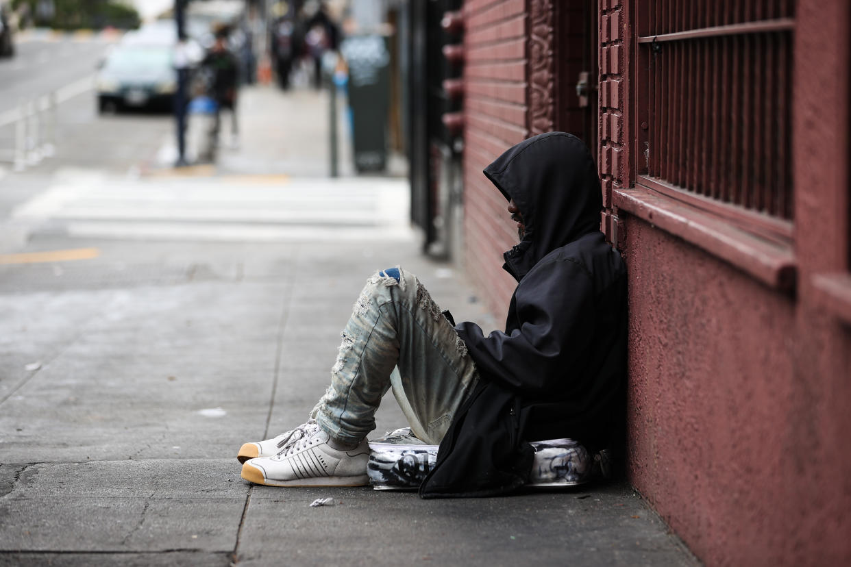 A person wearing a jacket with the hood up sits on the sidewalk with his back against a building.