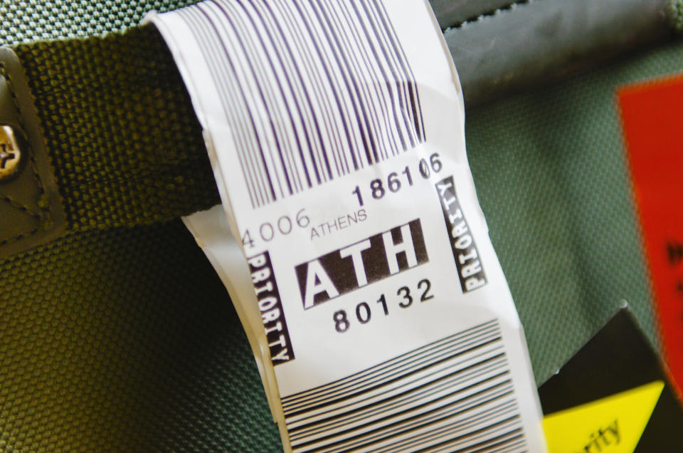 Luggage tag with the code "ATH" for Athens attached to a suitcase handle. The tag includes a barcode and priority markings