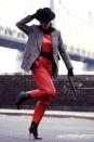 <p>A model wears a red jumpsuit, gray blazer, and black hat for photographs in front of a bridge in New York.</p>