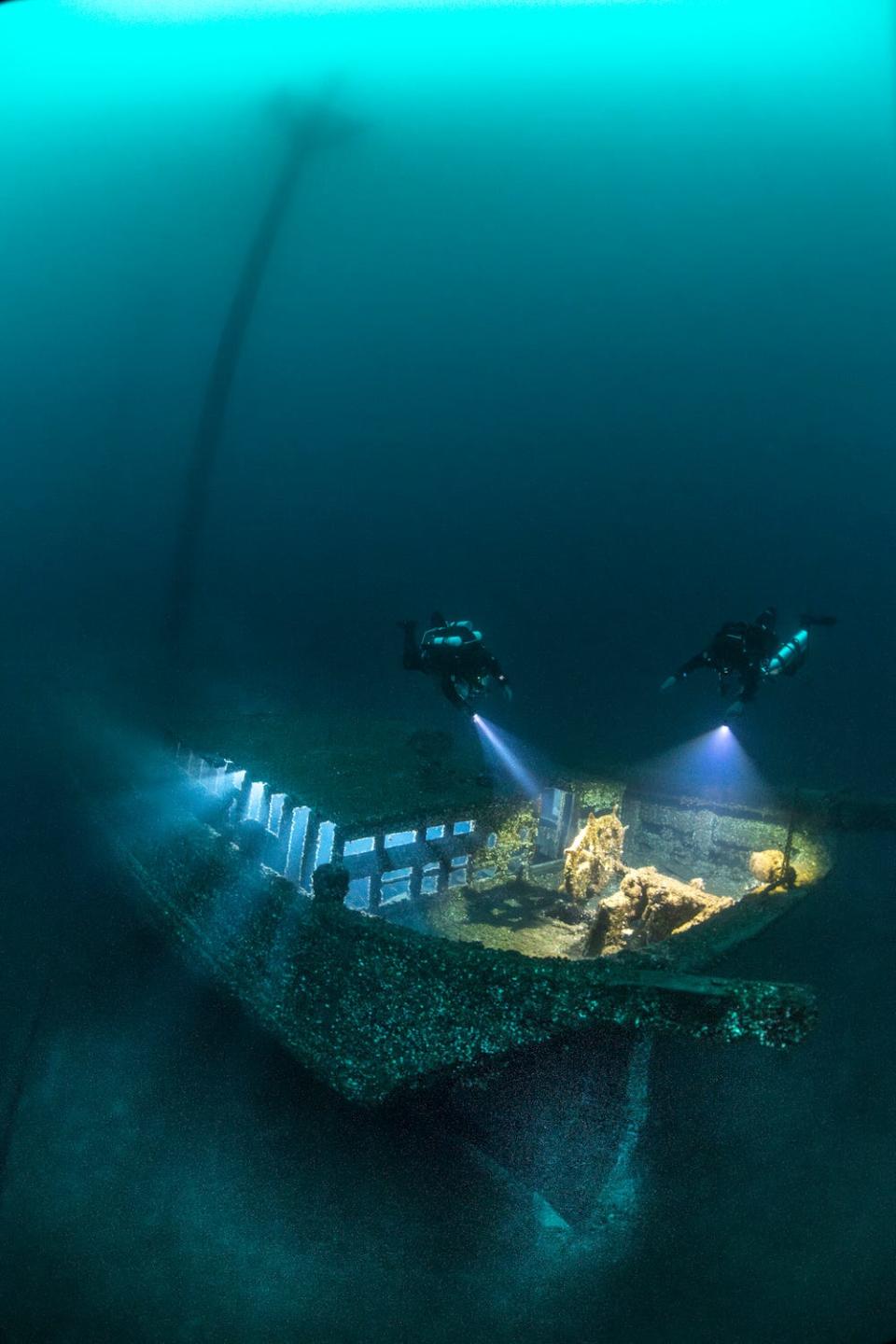 Two divers explore the wreck of the wooden schooner Kyle Spangler in Lake Huron, illuminating its deck with their torches. The image is a winner in the 2024 Underwater Photographer of the Year Awards.