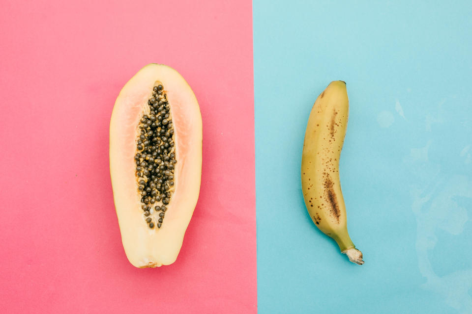 A halved papaya with seeds and a ripe banana are placed side by side on split pink and blue background