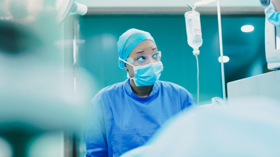 Woman adult black female nurse OR staff surgeon doctor talking discussing and looking at monitor vital signs with colleague standing at operating table patient while dressed in safety gown scrubs and performing operation in operating room medical facility hospital with staff team members support healthcare.