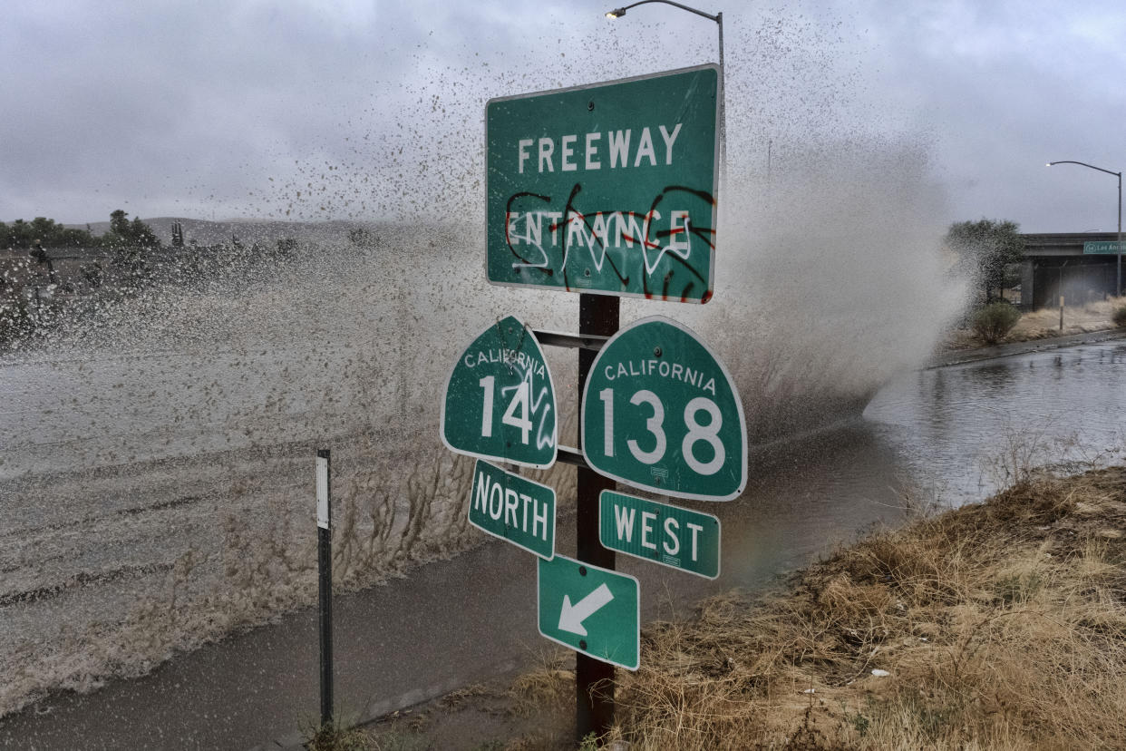 A large section of splashing water that appears to be caused by a speeding car erupts along a water-filled roadway next to a road sign that reads: Freeway entrance, California 14 north, California 138 west.