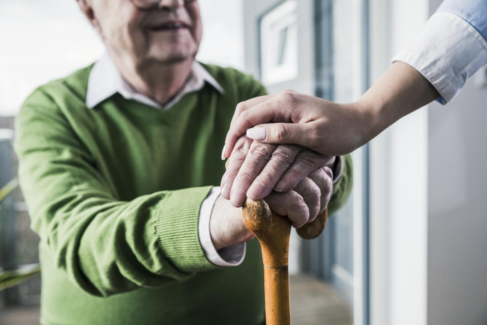 An elderly man with glasses holds a wooden cane while a younger person's hand rests reassuringly on his