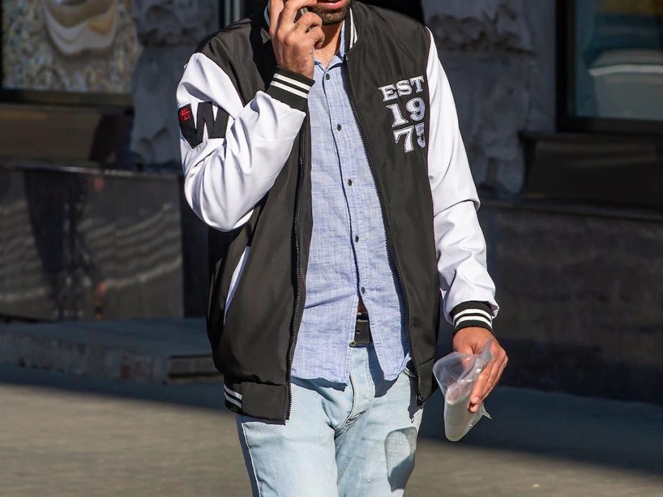 man walking down a street wearing a varsity jacket and jeans while talking on the phone