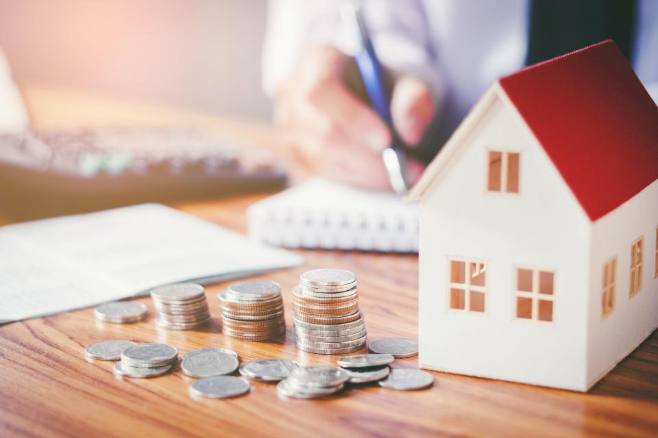 A toy house and three stacks of coins on a table with someone writing in a pad in the background.