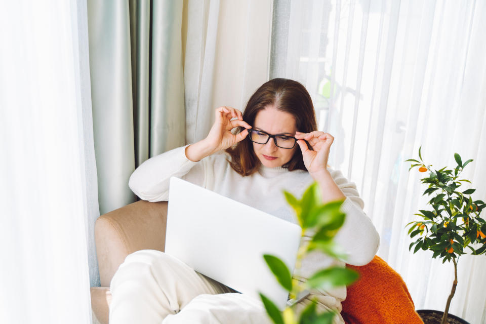 An individual with glasses and a white sweater sitting comfortably on a chair, adjusting their glasses while using a laptop, next to a potted plant