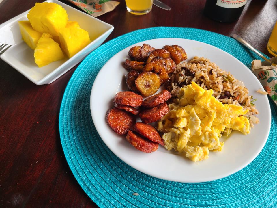 A white plate with a typical Costa Rican breakfast: eggs, gallo pinto, sausage, plantains, and pineapple.