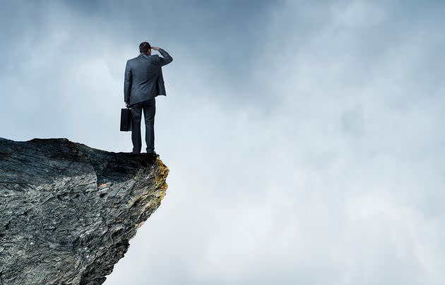 A businessman standing on the edge of a cliff and holding his briefcase, peers out towards a fog shrouded sea.  Ample copy space is created by the clouds.