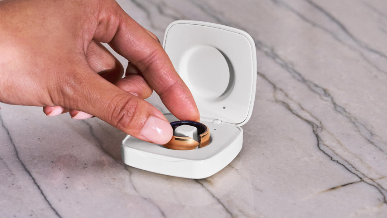  The Evie smart ring in its charging case, with a woman's hand reaching to pick it up. . 