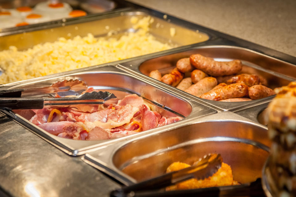 A buffet featuring scrambled eggs, sausages, bacon, and hash browns. Tongs are provided for self-service