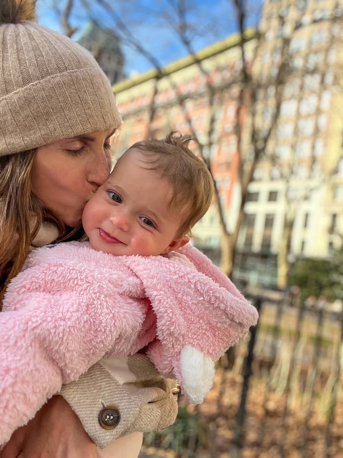 Woman kissing baby on forehead, both in warm attire, outdoors with blurred trees and buildings in background