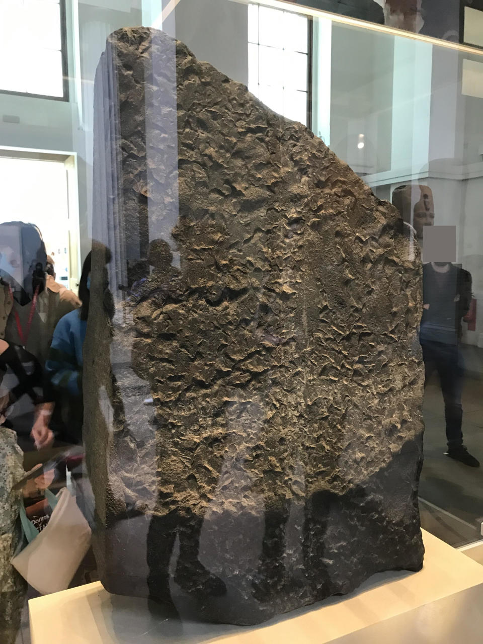 The huge stone's uneven, broken surface, behind glass, with people standing next to it