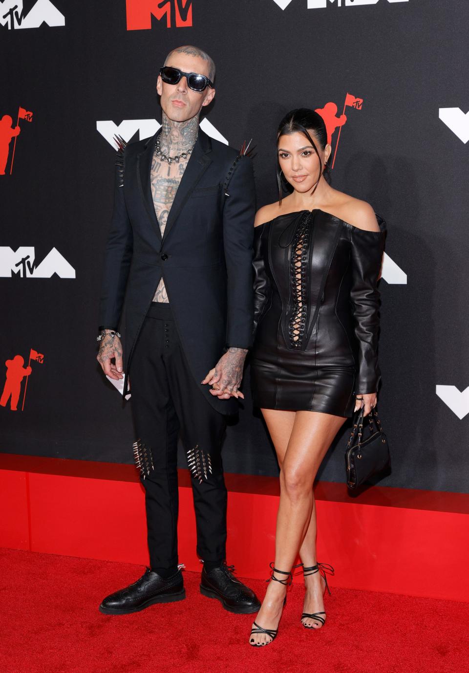 Travis next to Kourtney in a black off the shoulder leather minidress with long sleeves and a lace up neckline from her belly button.