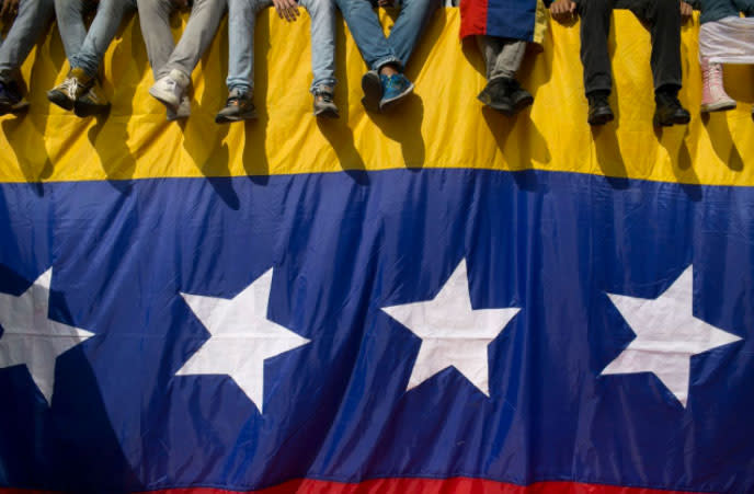 In Venezuela protestieren die Menschen gegen den Präsidenten Nicolás Maduro. In Caracas sitzen einige Personen auf einer Mauer, über die die Landesflagge gespannt ist. Die Demonstranten wollen, dass Maduro sein Amt niederlegt. (Foto: Alejandro Cegarra/AP)