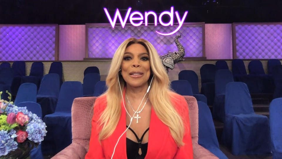 Wendy Williams in a bright orange pink jacket sitting alone in the audience of her show with headphones on