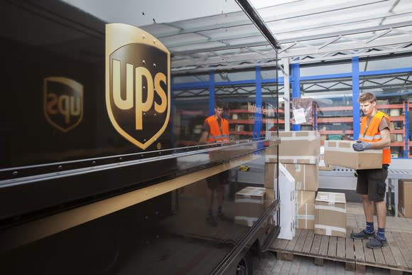 A male UPS employee wearing brown shorts and a neon orange vest loading boxes onto a truck.